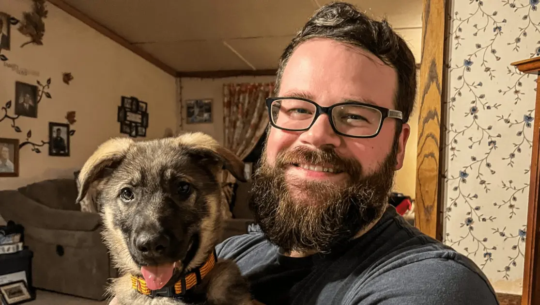 A picture of Deputy Editor Aaron Gates, a bearded man with dark hair and glasses, who is smiling while holding a German Shepherd puppy.