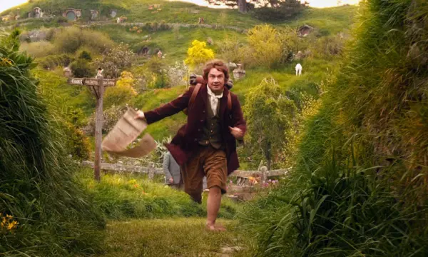 Bilbo Baggins, a hobbit with a backpack and a long piece of parchment, runs through green hills.